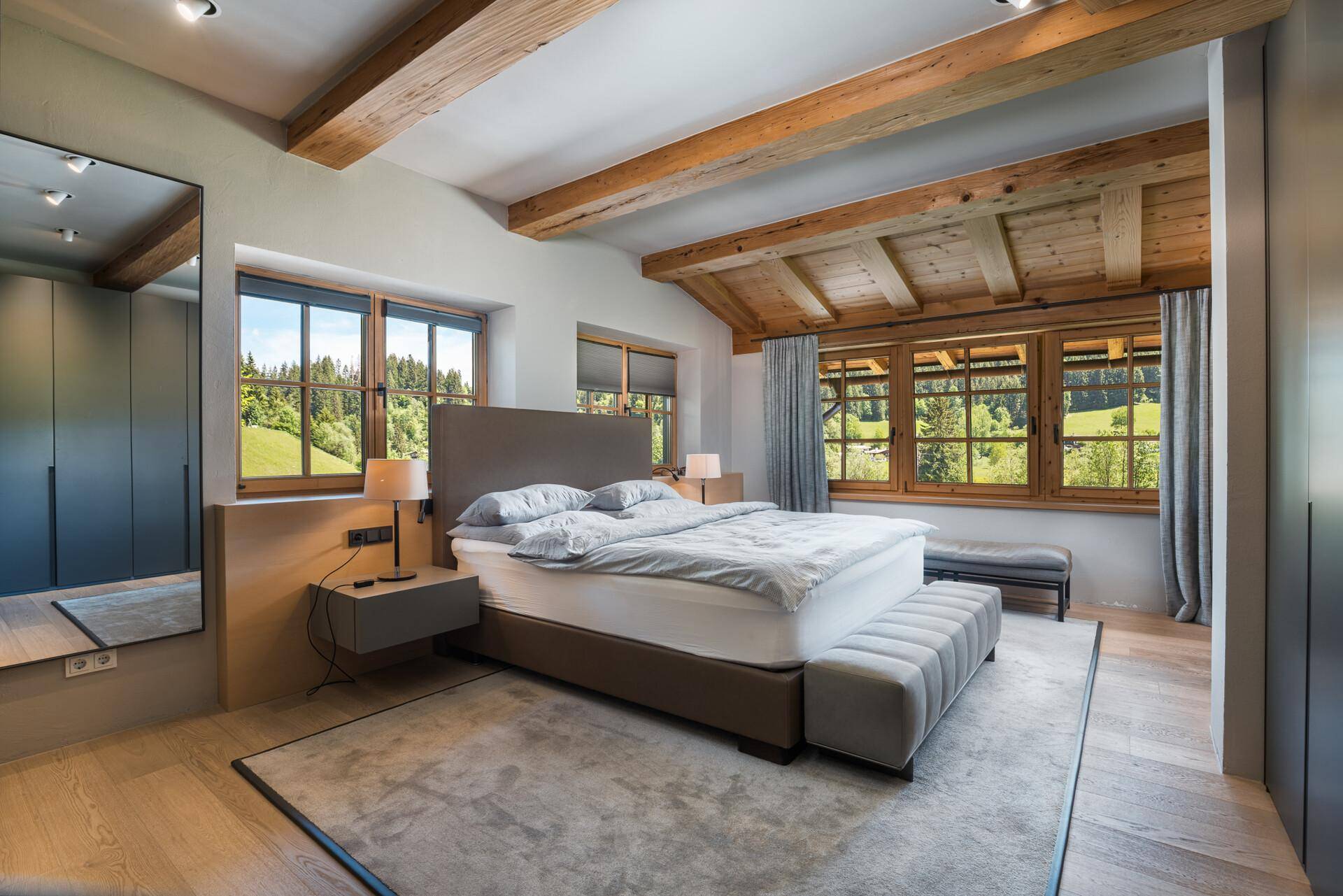 Gemütliches Schlafzimmer mit Holzbalkendecke, großen Fenstern und Blick auf die grüne Landschaft.