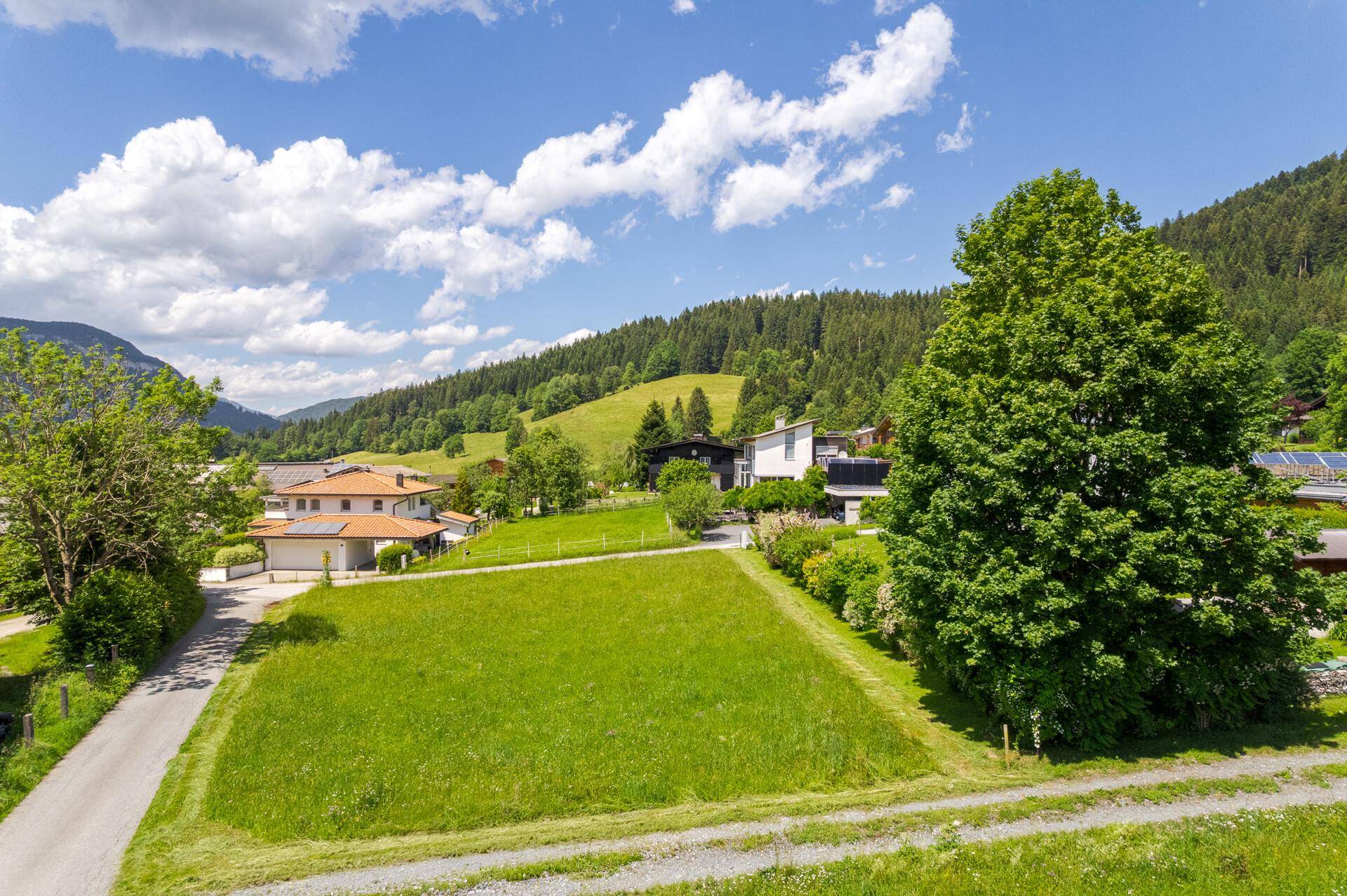 Blick über grüne Wiesen auf eine ländliche Siedlung, umrahmt von bewaldeten Hügeln unter blauem Himmel.