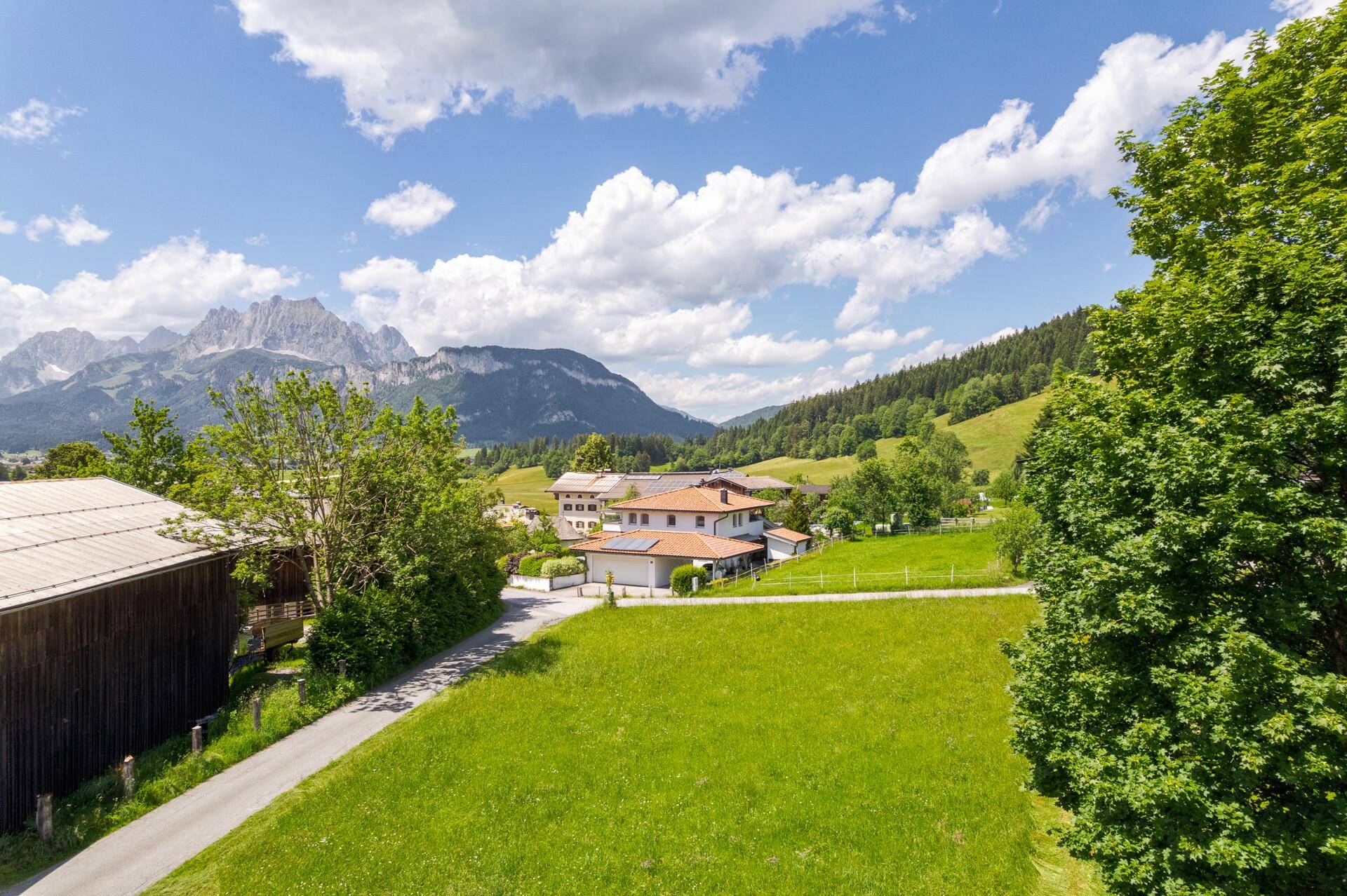 Sonnige Alpenlandschaft mit grünen Feldern, traditionellen Häusern und imposanten Berggipfeln am Horizont.