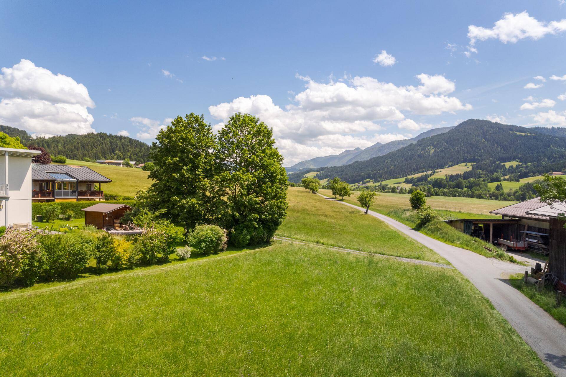 Panoramablick über grüne Felder und Hügel, mit einem modernen Wohnhaus am Rand und Bergen in der Ferne.