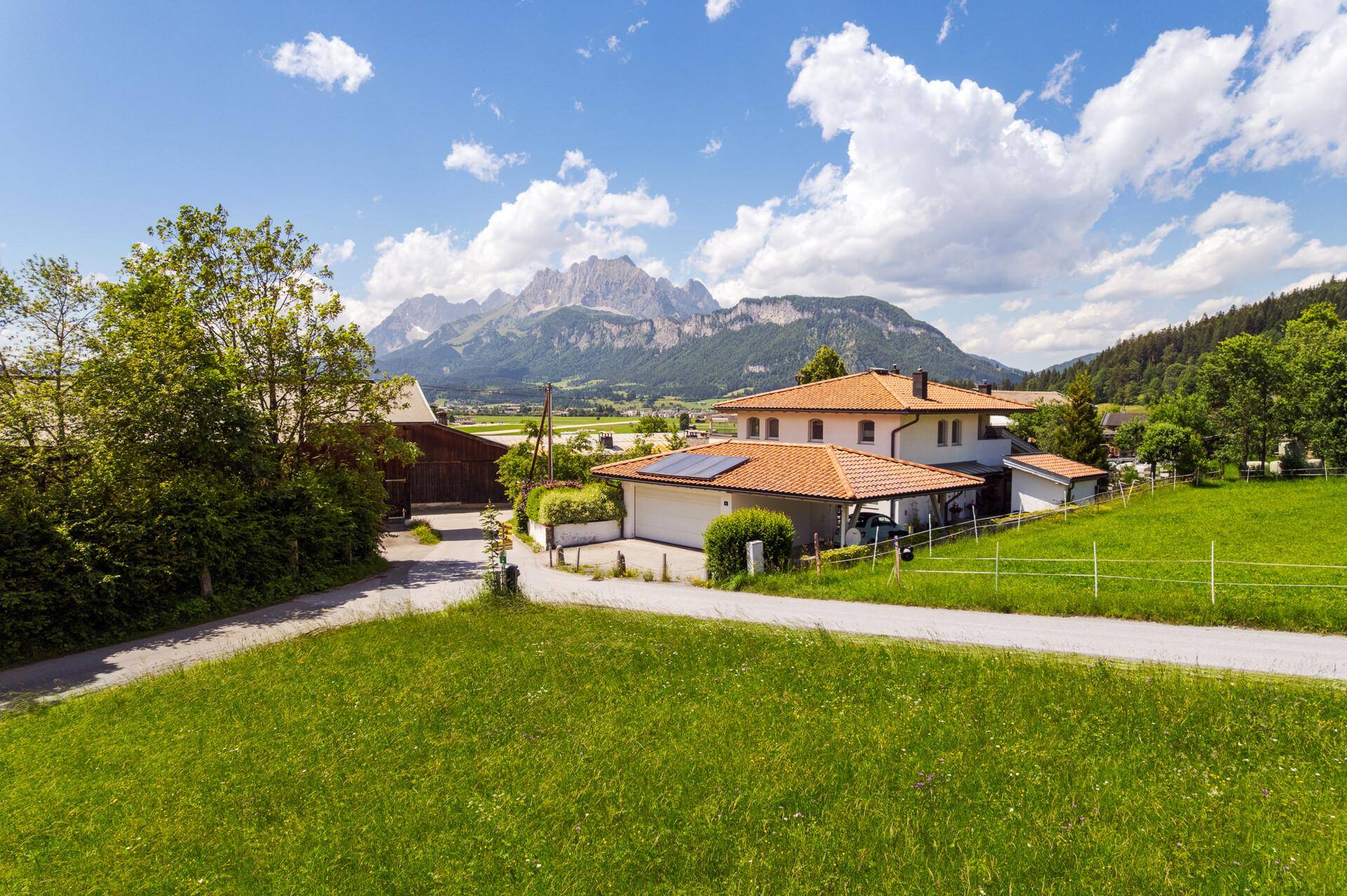 Idyllischer Ausblick auf grüne Felder, ein Wohnhaus mit Solaranlage und die beeindruckende Bergkulisse der Alpen.