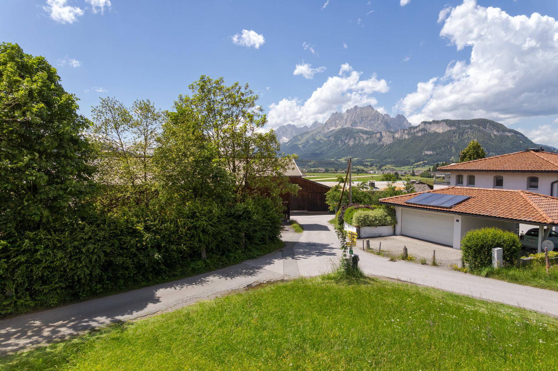Sonnige Landschaft mit grünen Wiesen, einem Wohnhaus mit Solaranlage und majestätischen Bergen im Hintergrund.
