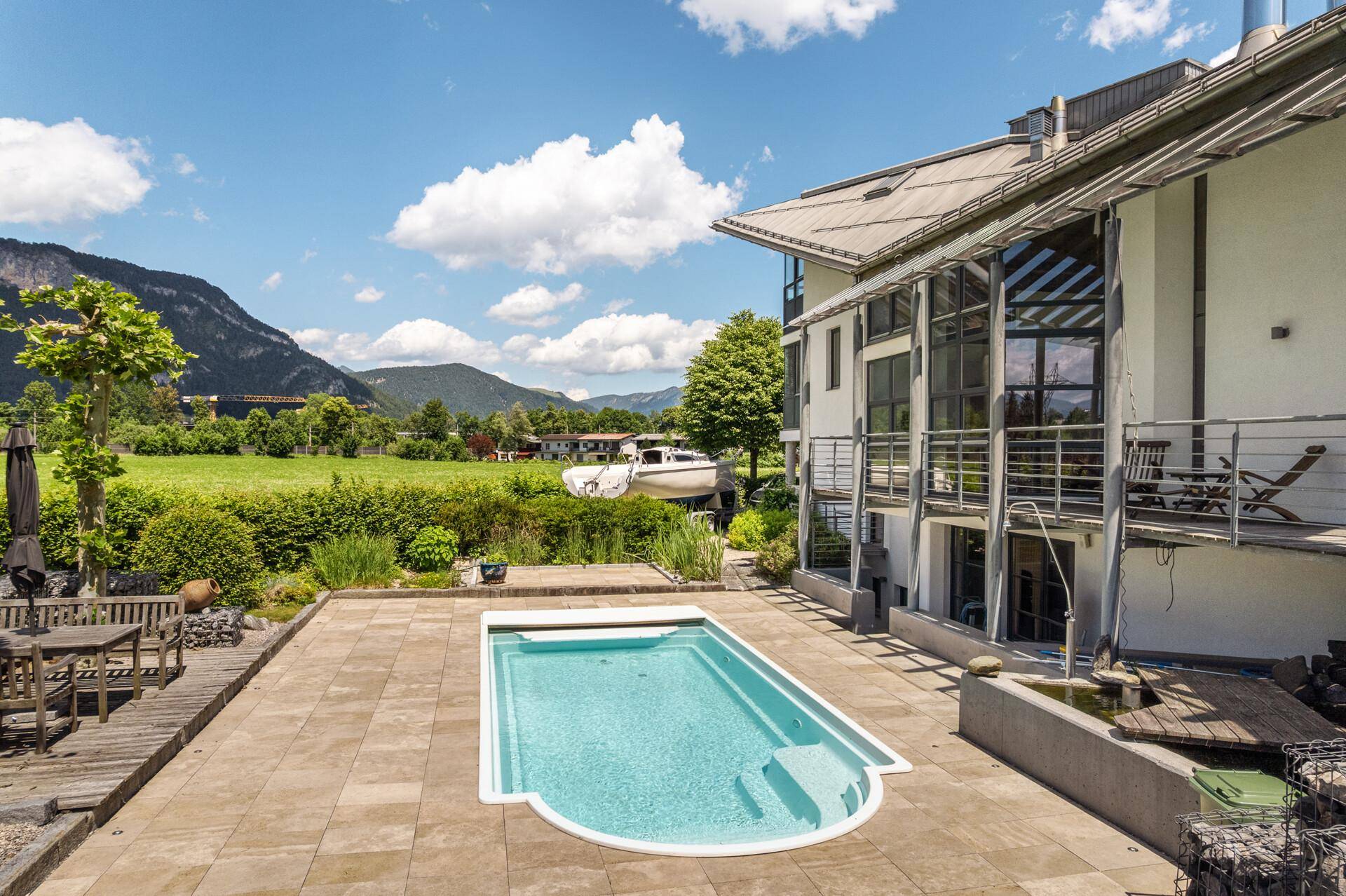 Außenbereich mit Swimmingpool, gepflasterter Fläche und Blick auf die moderne Hausfassade und Berge.