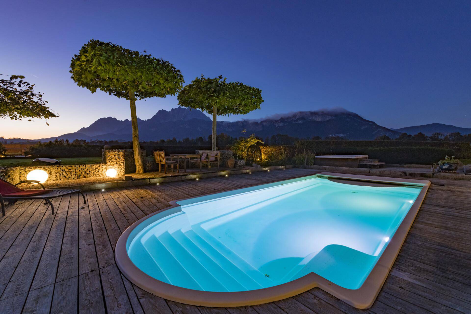 Beleuchteter Poolbereich mit Holzdeck und Bergblick in der Abenddämmerung, umgeben von Bäumen.