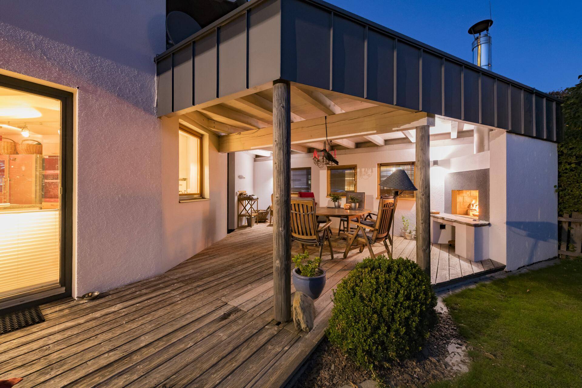 Gemütliche überdachte Holzterrasse mit Essbereich und integriertem Außenkamin am Abend.