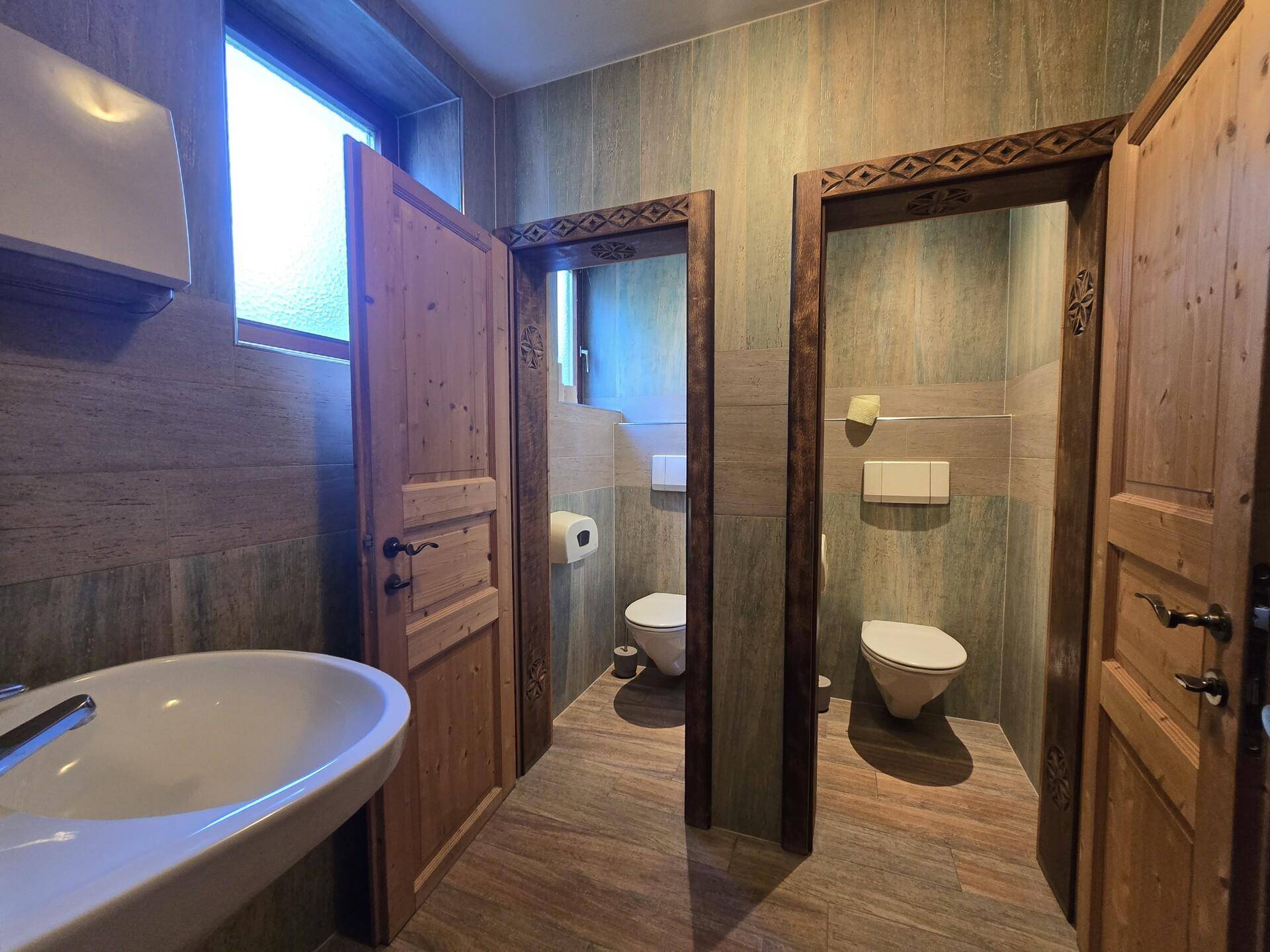 Mehrere Toilettenkabinen in einem öffentlichen Waschraum mit gefliesten Wänden und Holztüren.