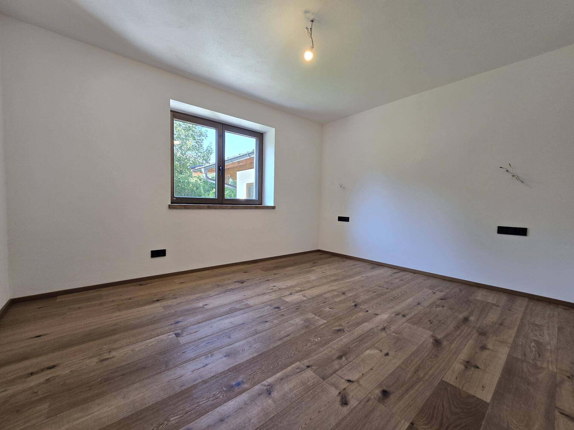 Helles Schlafzimmer mit Eichenparkett und einem Fenster mit Blick ins Grüne.