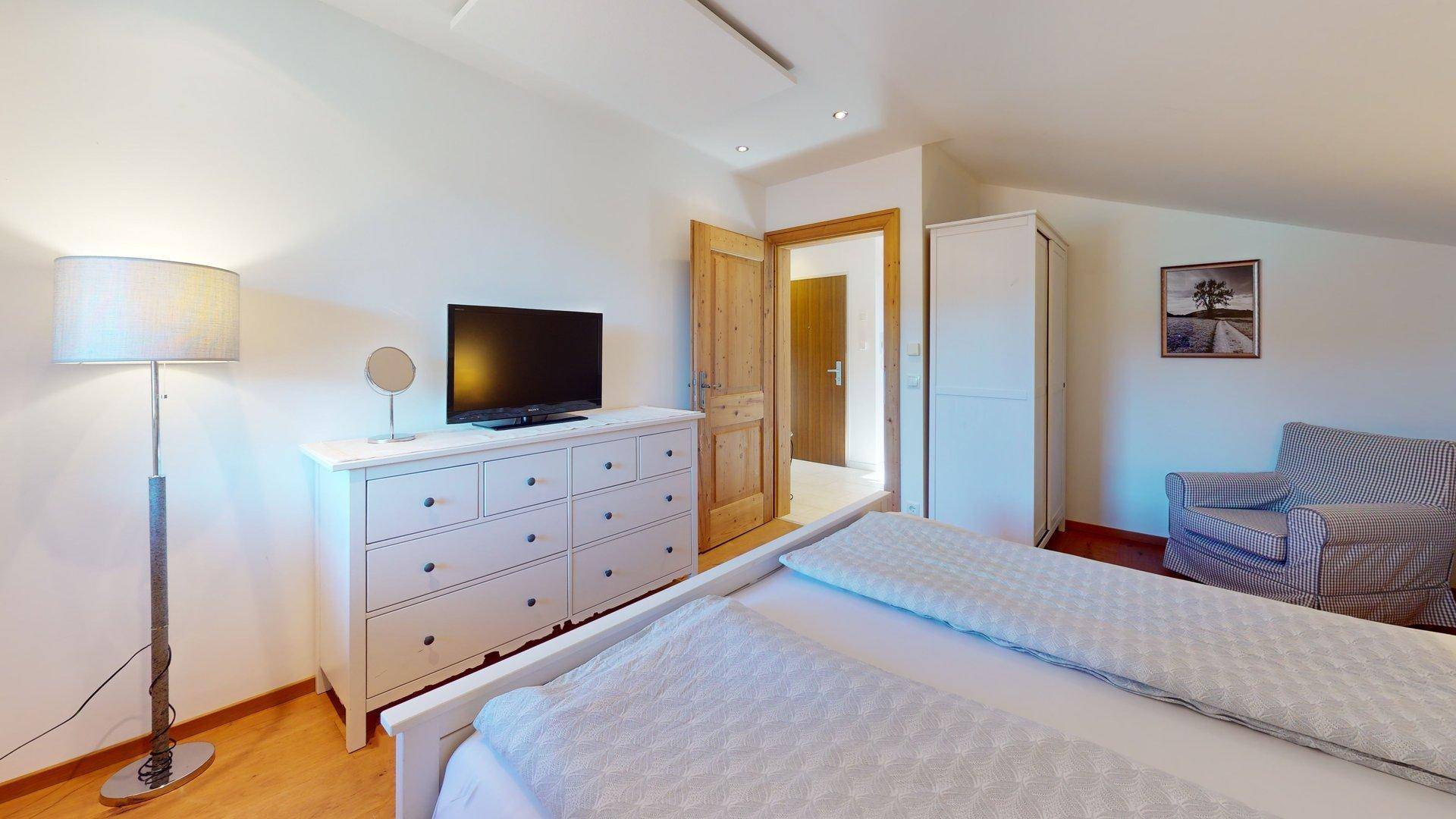 Helles Schlafzimmer mit Doppelbett, Kommode und Stehlampe, Zugang zu weiterem Raum.