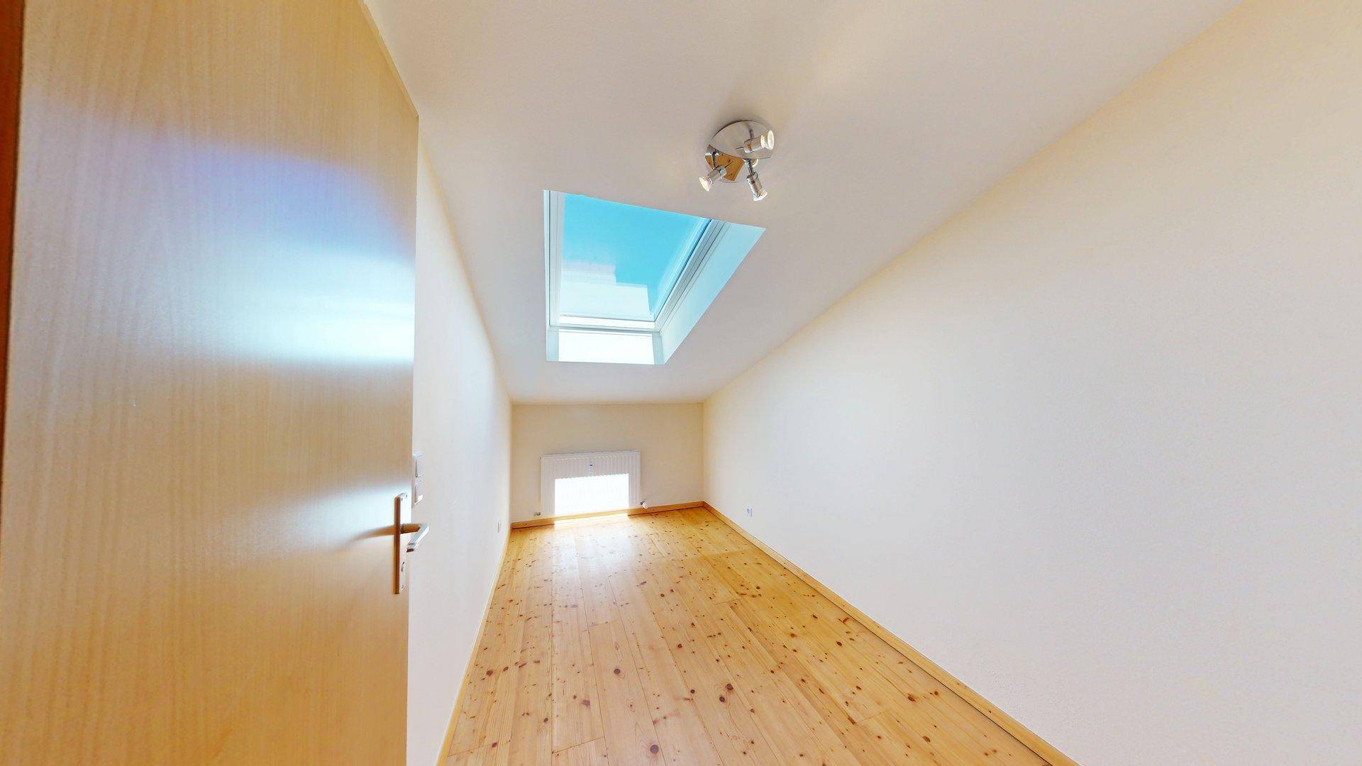 Schmaler Raum im Dachgeschoss mit Holzboden, Dachfenster und natürlichem Lichteinfall.
