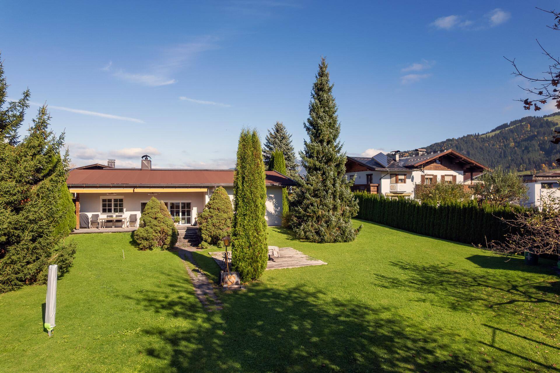 Sonniger Garten mit weitläufiger Rasenfläche, hohen Bäumen und einem charmanten Einfamilienhaus im Hintergrund.