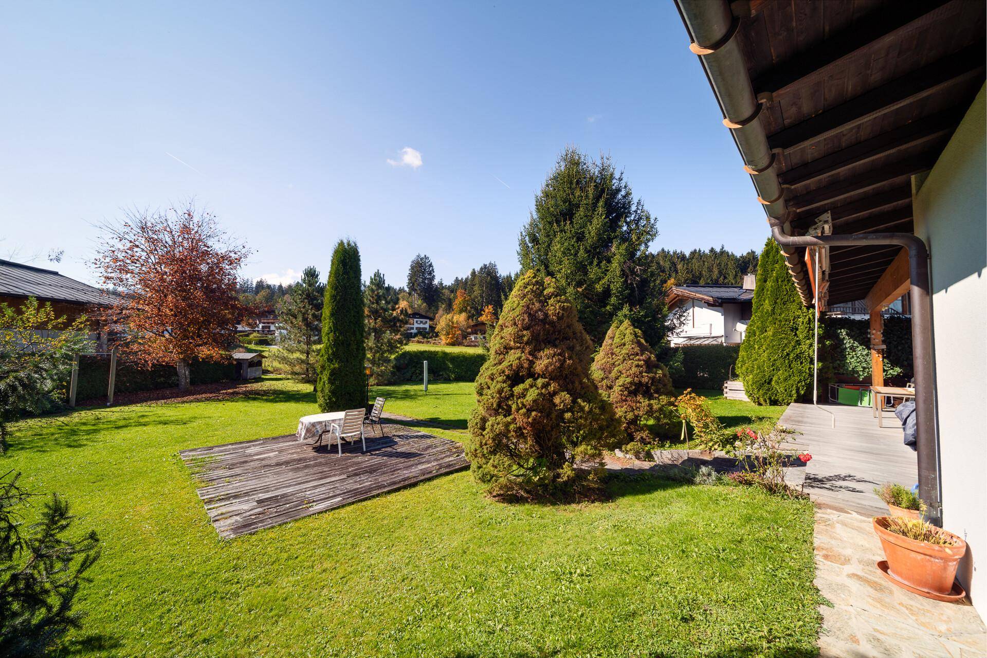 Großer Garten mit Holzterrasse und Sitzgelegenheit, umgeben von Bäumen und Sträuchern in herbstlicher Färbung.