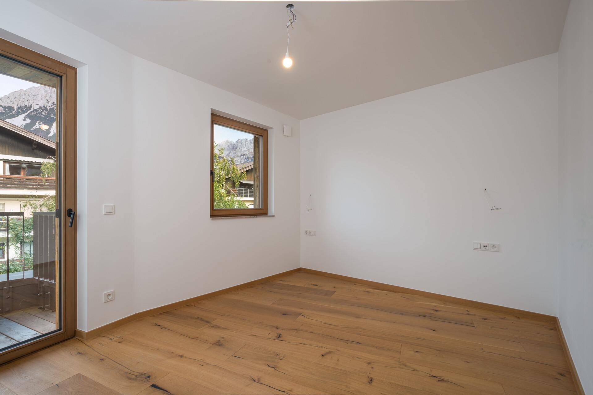 Helles, leeres Zimmer mit Holzboden, Fenster und Zugang zu einem Balkon mit Bergblick.