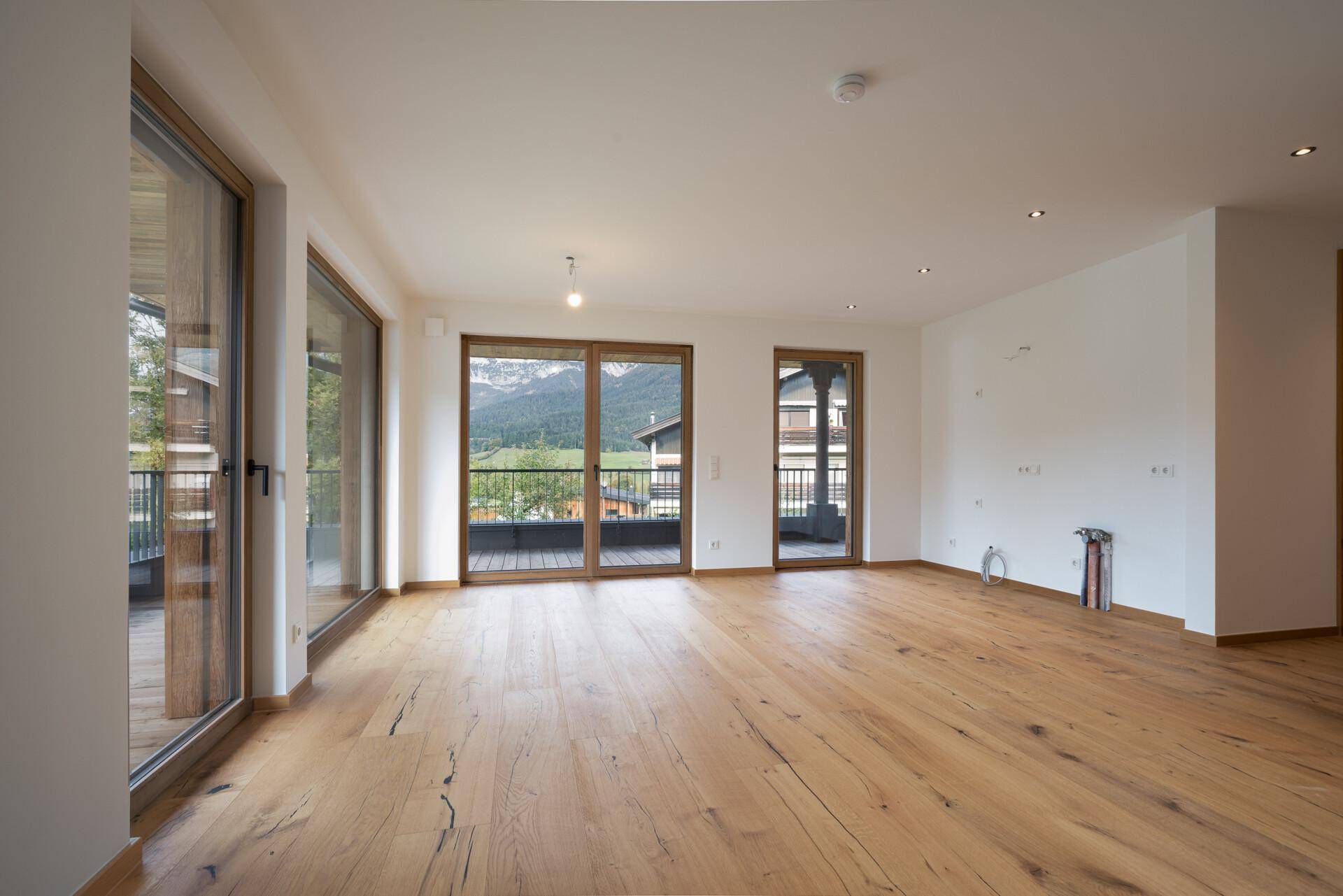 Geräumiger, unmöblierter Wohnbereich mit Holzboden und großen Fenstern mit Bergblick.
