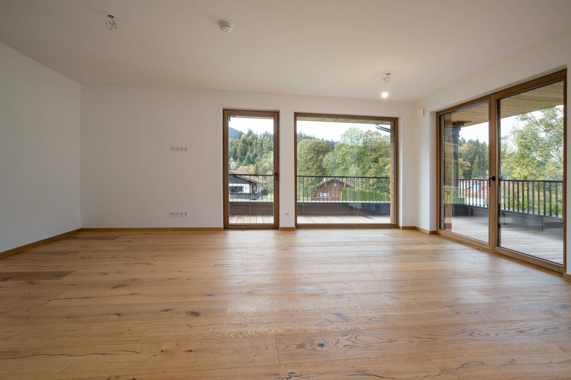 Heller, leerer Wohnraum mit Holzboden und großen Fenstern, die einen grünen Ausblick bieten.
