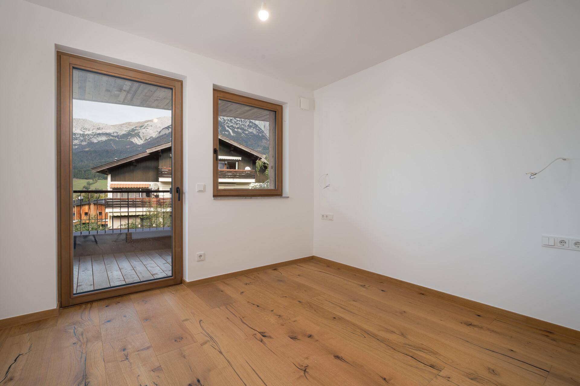Unmöbliertes Zimmer mit Holzboden, Fenster und Balkonzugang, ideal als Kinderzimmer mit Bergblick.