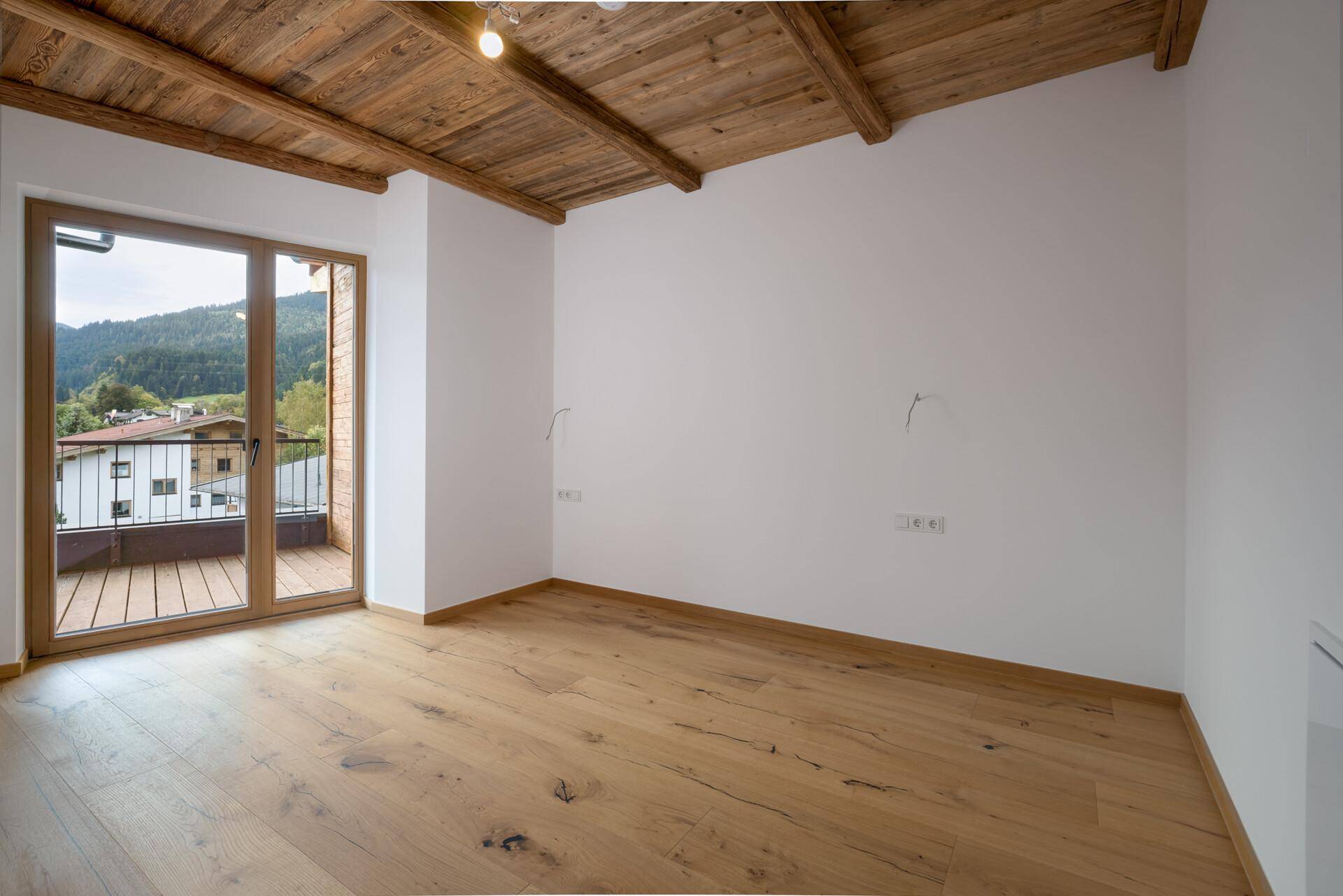 Helles, leeres Zimmer mit Holzboden, Holzbalkendecke und direktem Zugang zu einem Balkon.