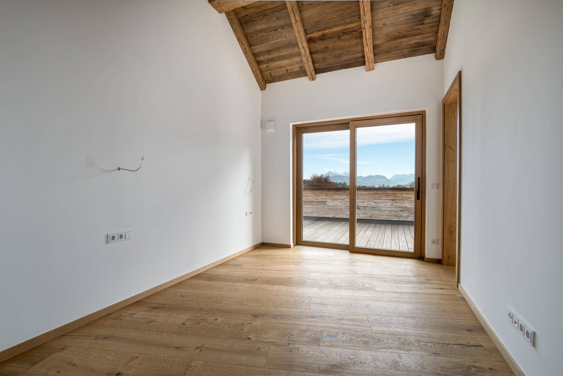 Helles, leeres Zimmer mit Holzboden, Holzbalkendecke und Schiebetür zu einer Terrasse mit Bergblick.