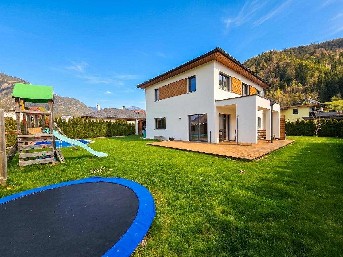 Hinterhof des Hauses mit großer Rasenfläche, Spielplatz und Trampolin, umgeben von Bergen.