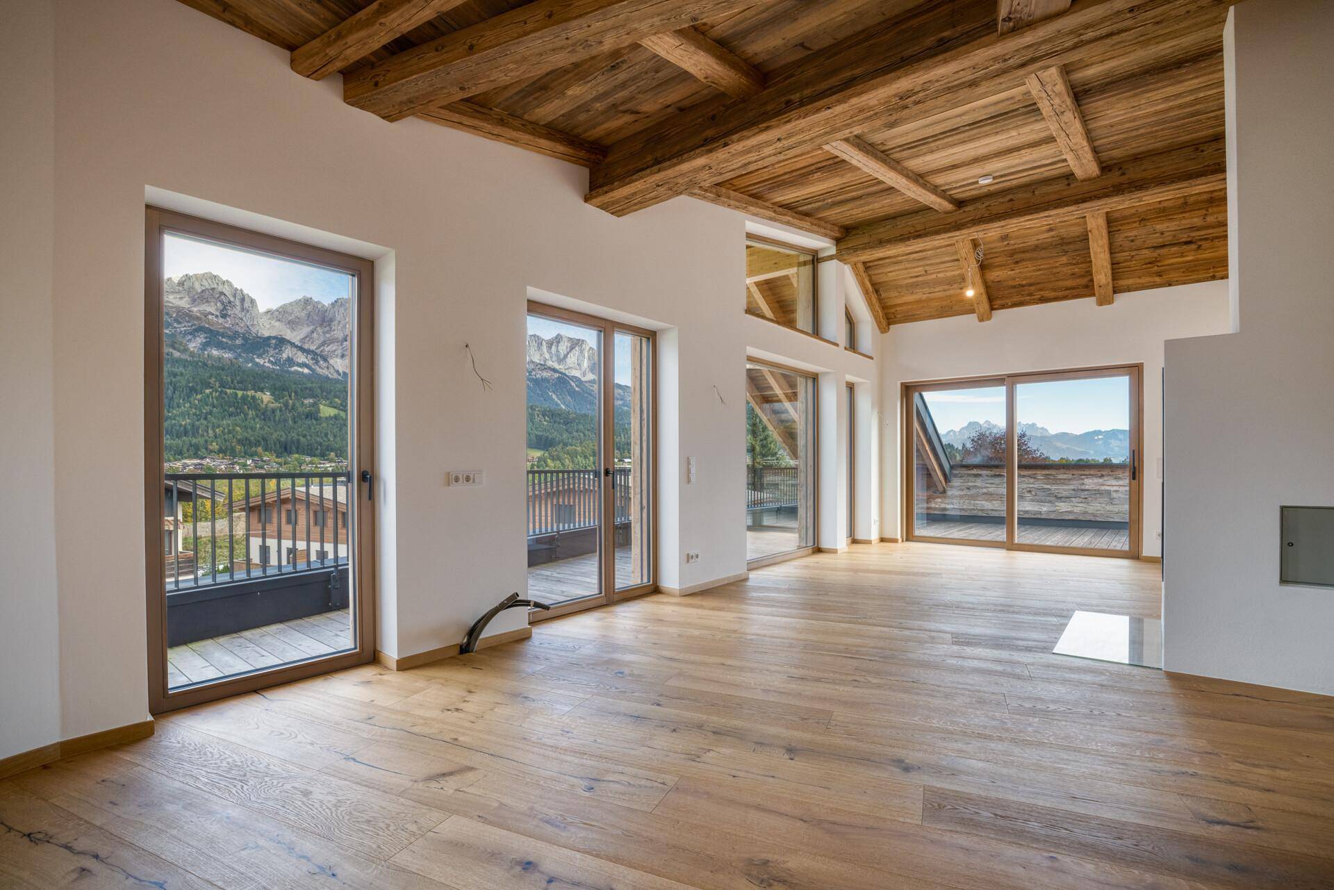 Heller, leerer Wohnbereich mit Holzboden, Holzbalkendecke und großen Fenstern mit Bergblick.