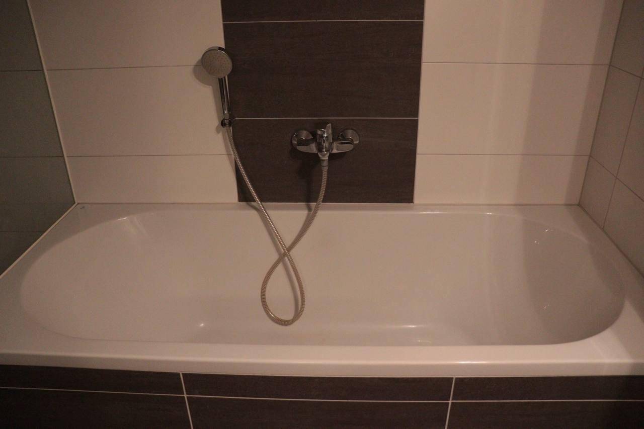 Nahaufnahme einer Badewanne mit integrierter Handbrause, umgeben von modernen Fliesen.