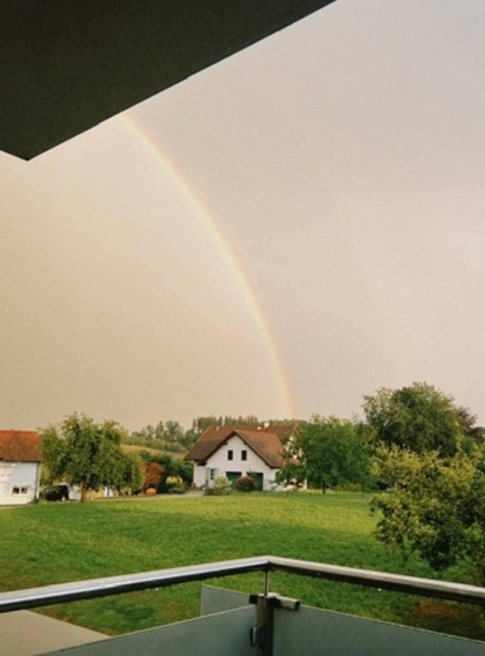 Malerischer Ausblick vom Balkon auf eine grüne Landschaft mit Häusern und einem Regenbogen am Himmel.