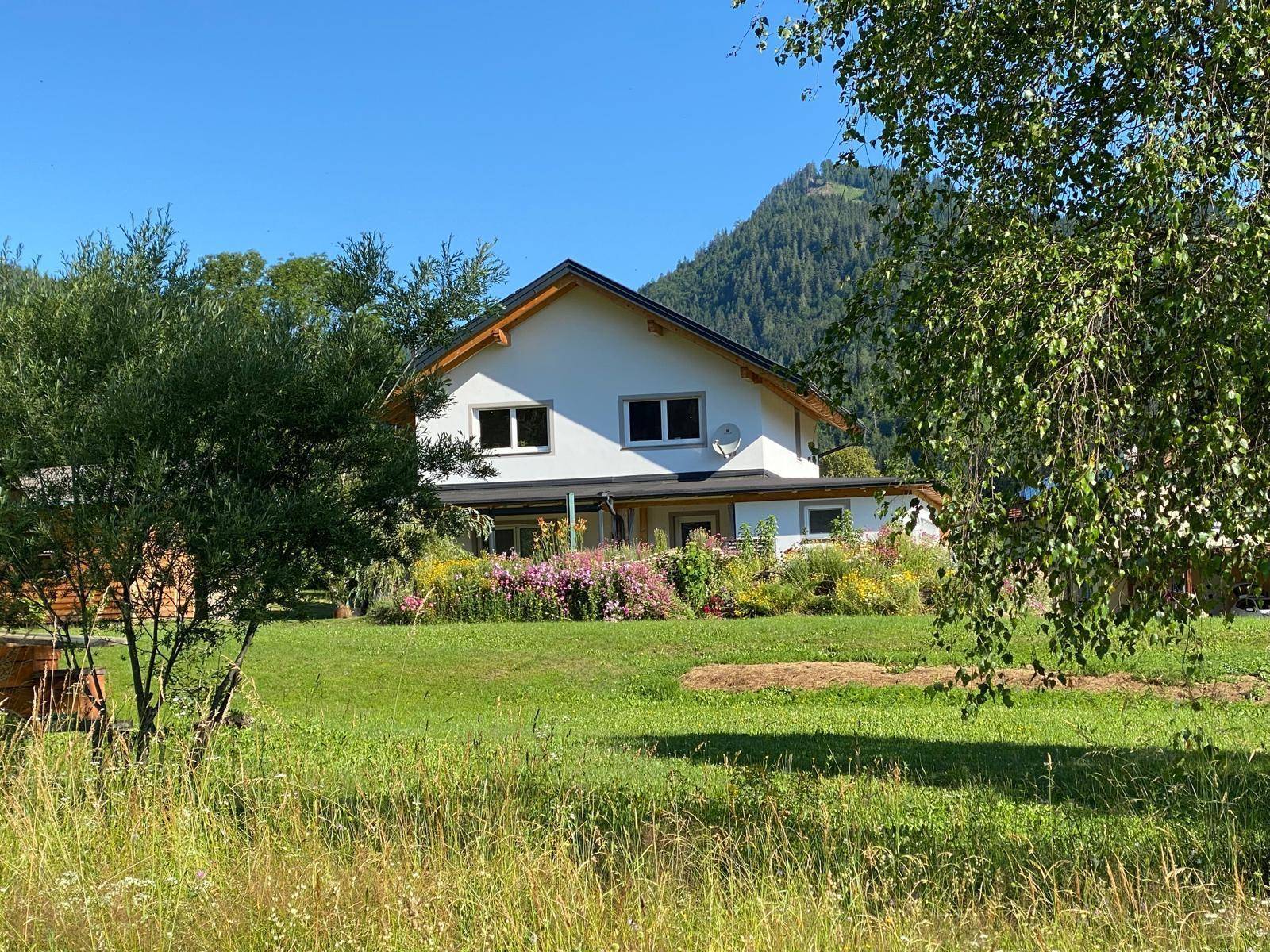Rückseite des Hauses mit blühenden Blumenbeeten und weitläufigem Rasen vor Bergkulisse.