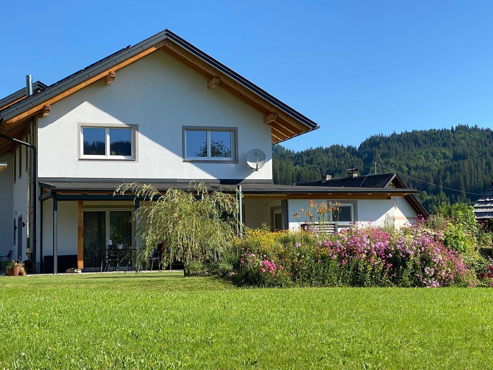 Hausfassade mit blühenden Blumenbeeten und weitem Blick auf die umliegende Natur.
