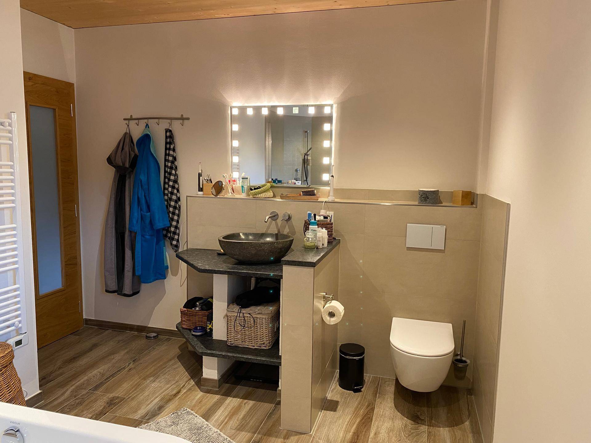 Helles Badezimmer mit WC, Steinwaschbecken und großem beleuchteten Spiegel.