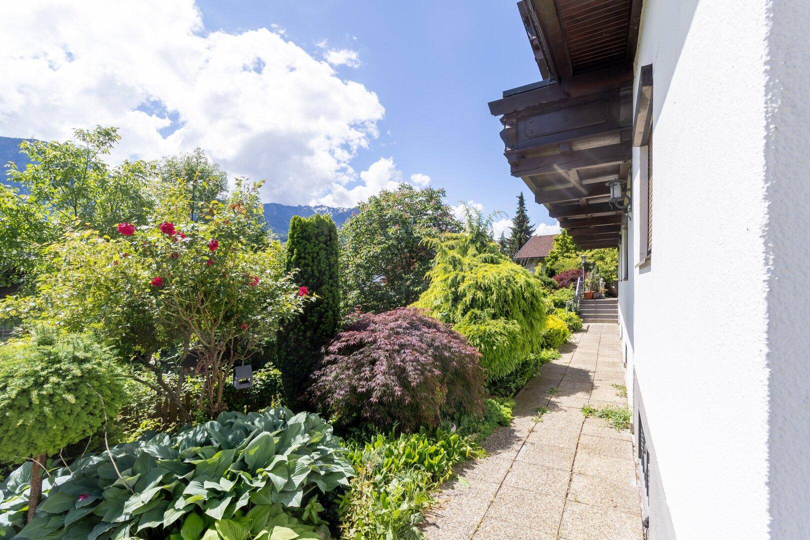 Gartenweg entlang des Hauses, umgeben von vielfältiger Bepflanzung und Bäumen unter blauem Himmel.