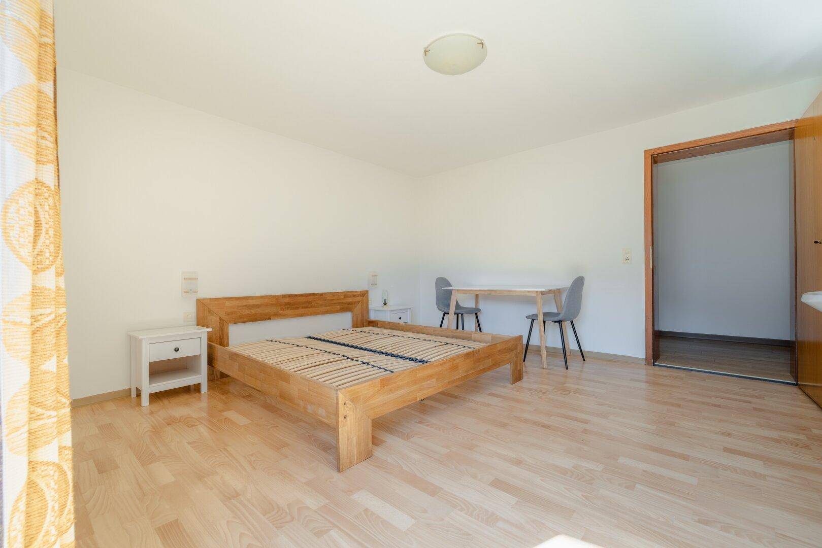 Helles Schlafzimmer mit Holzbett, Laminatboden und einem kleinen Tisch, bietet eine ruhige Atmosphäre.