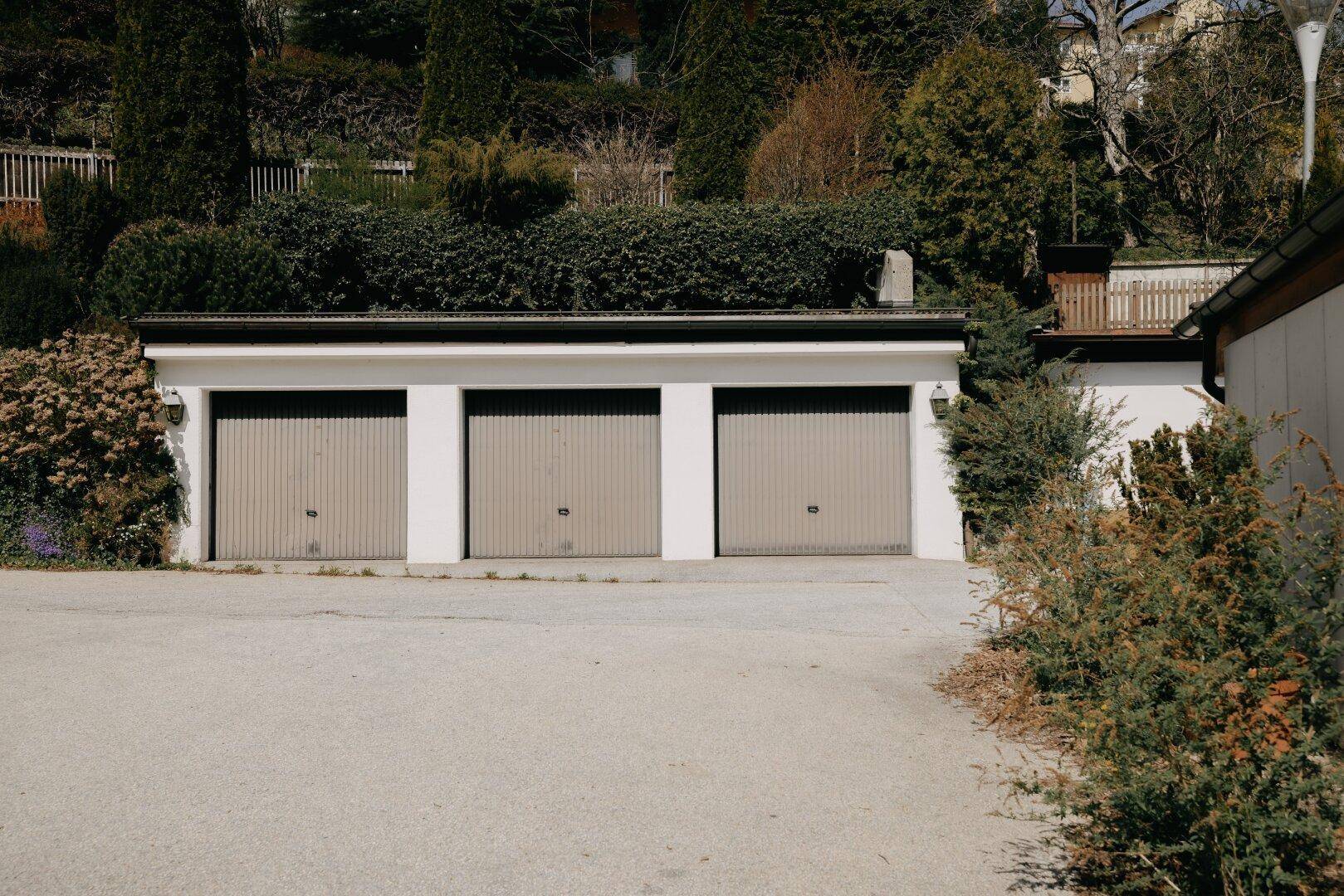 Drei Garagen mit hellen Toren, umgeben von grüner Bepflanzung und Zufahrt.