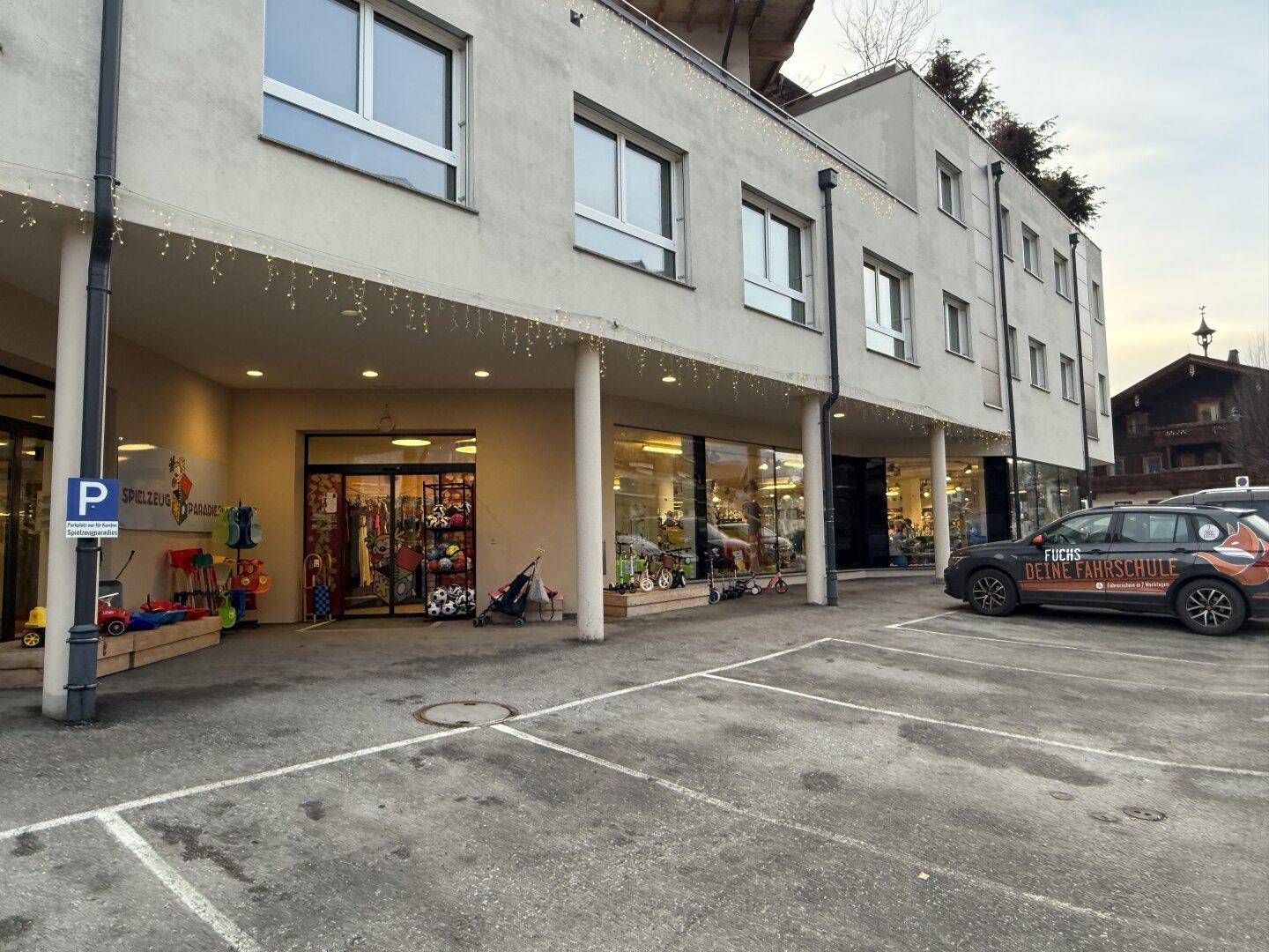 Erdgeschossfassade mit Schaufenstern, Eingangsbereich und einem gepflasterten Parkplatz.