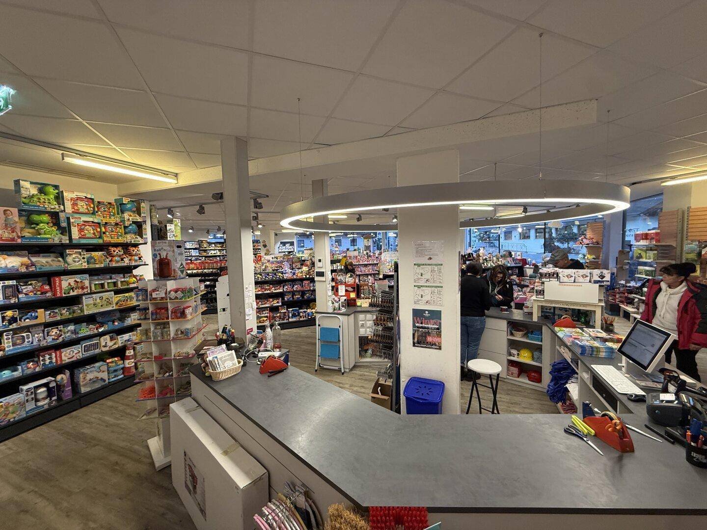 Kassenbereich eines Ladens mit Verkaufstheke und Kunden, umgeben von Produkten.