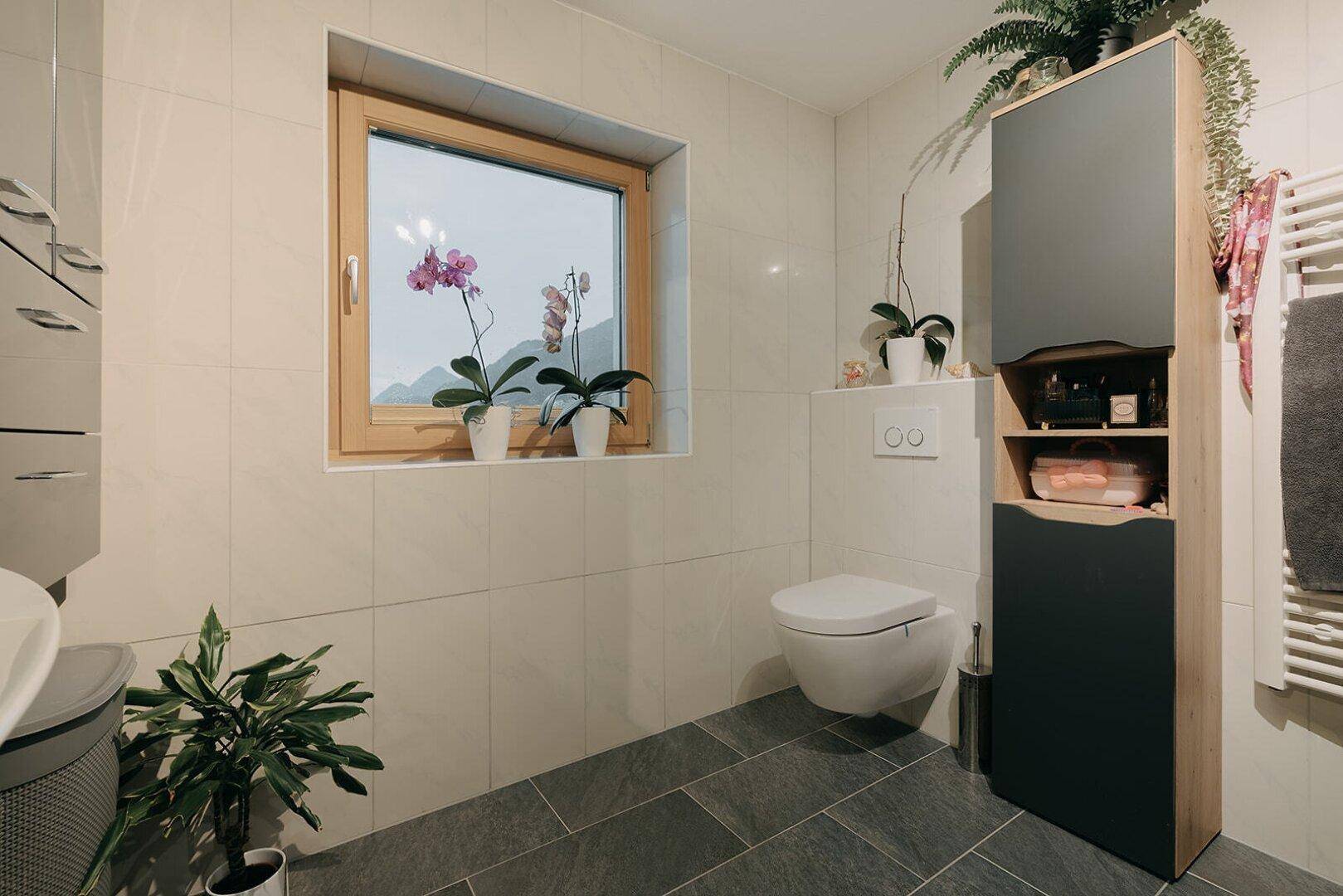 Modernes Badezimmer mit wandhängendem WC, Fenster und hohem Schrank für Stauraum.