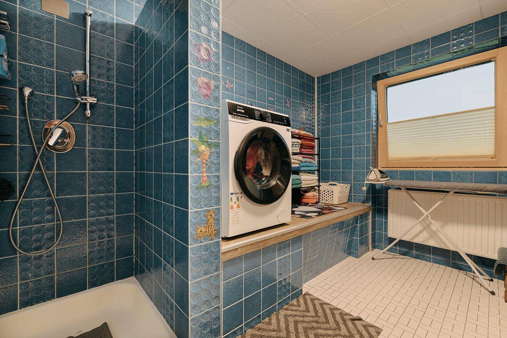 Waschraum mit blauen Fliesen, Dusche und Waschmaschine, gut beleuchtet durch ein Fenster.