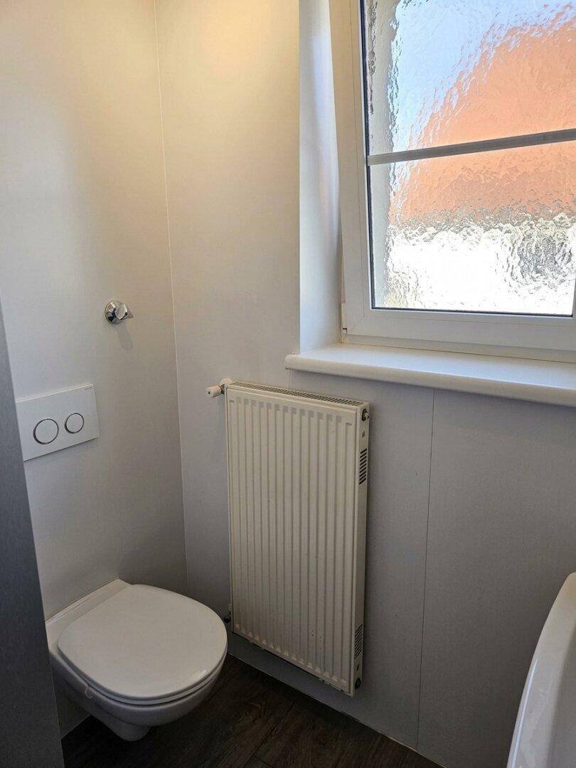 Separates WC mit wandhängender Toilette, Heizkörper und einem Fenster für natürliche Belüftung.
