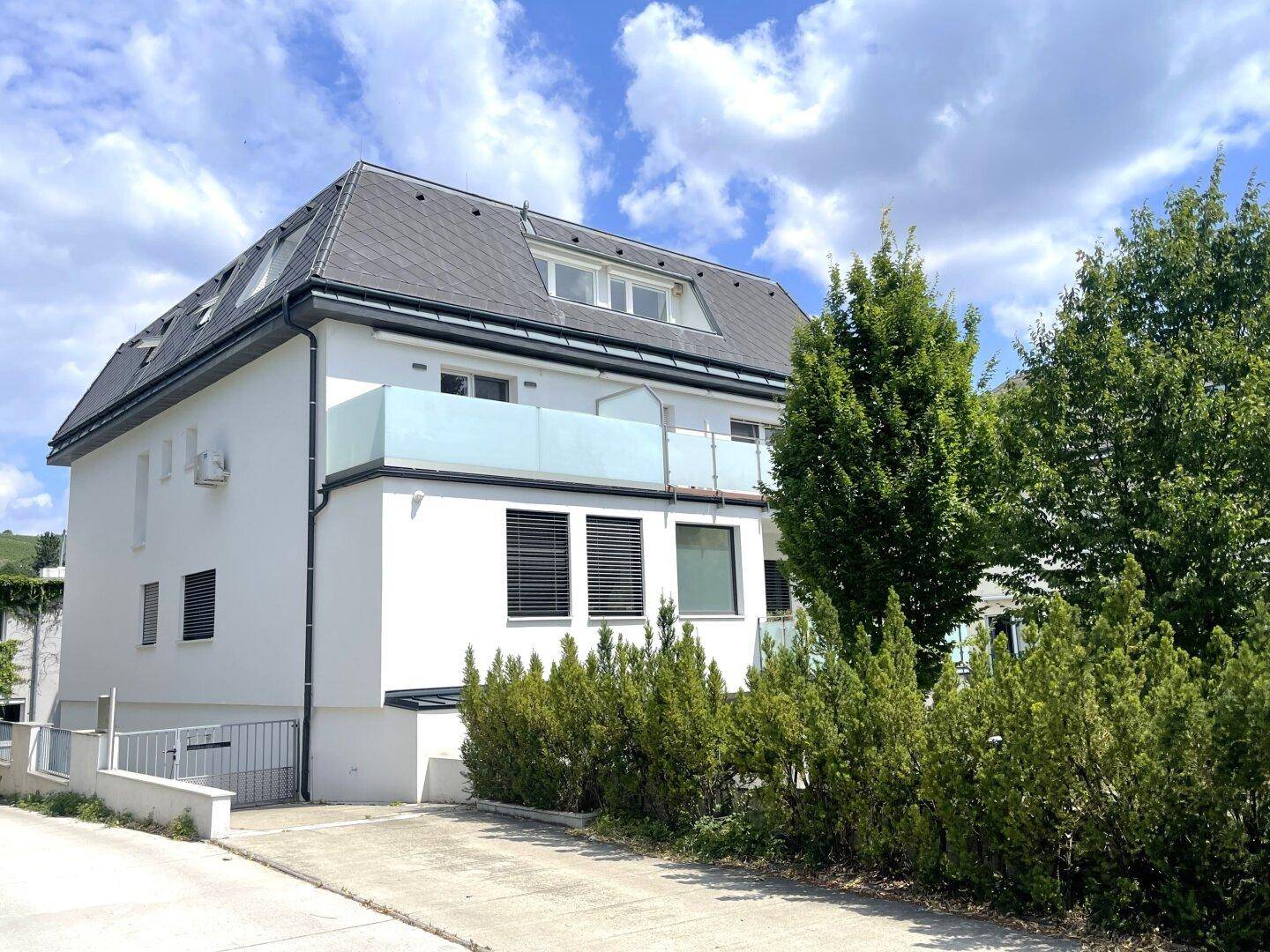 Modernes Wohnhaus mit weißer Fassade, Mansarddach und Balkonen mit Glasbrüstungen.