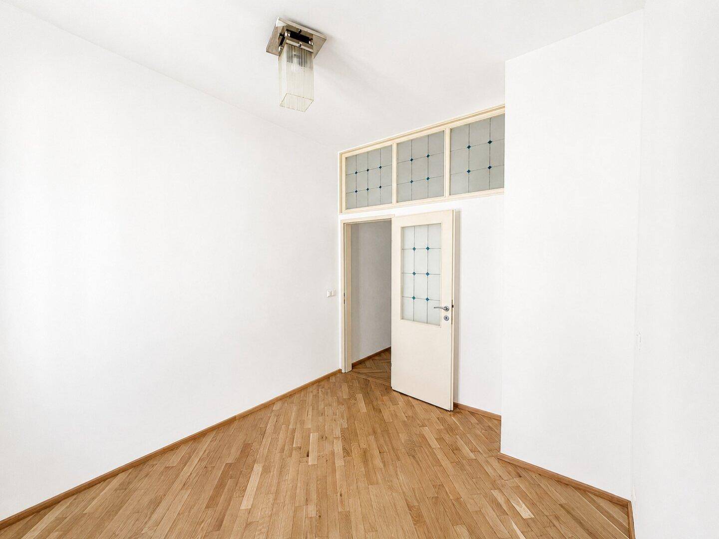Helles Zimmer mit Fischgrätparkett und einer Tür mit dekorativem Oberlicht in einem Altbau.