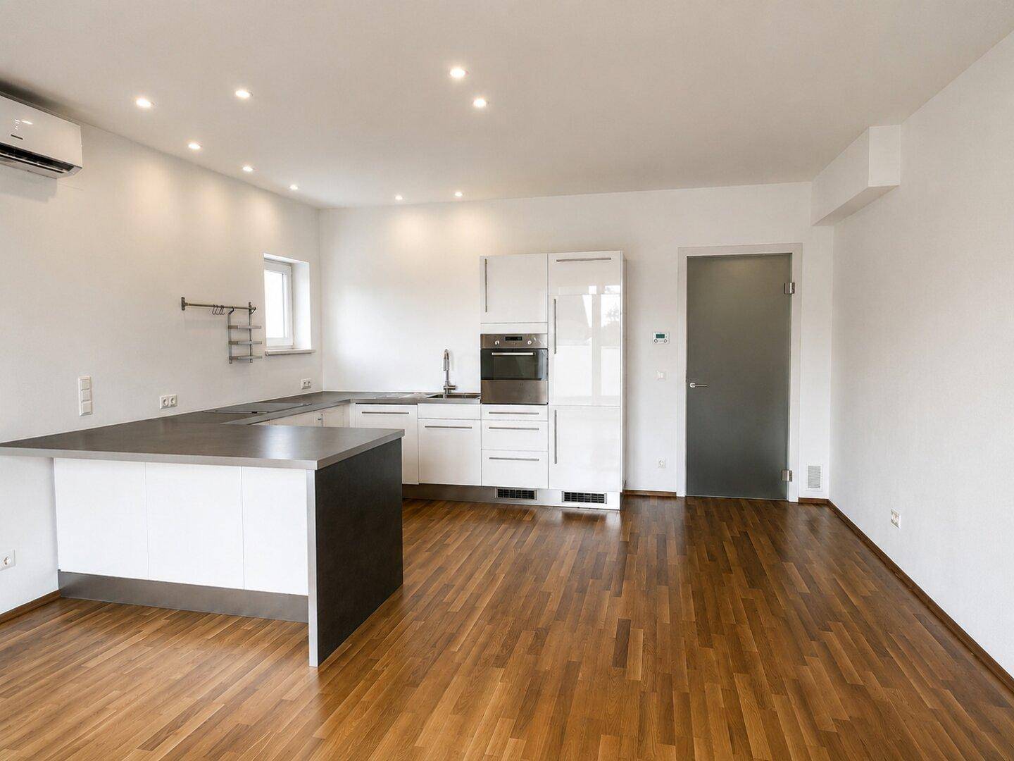 Moderne Einbauküche mit weißen Hochglanzfronten, dunkler Arbeitsplatte und Holzboden in offenem Wohnbereich.