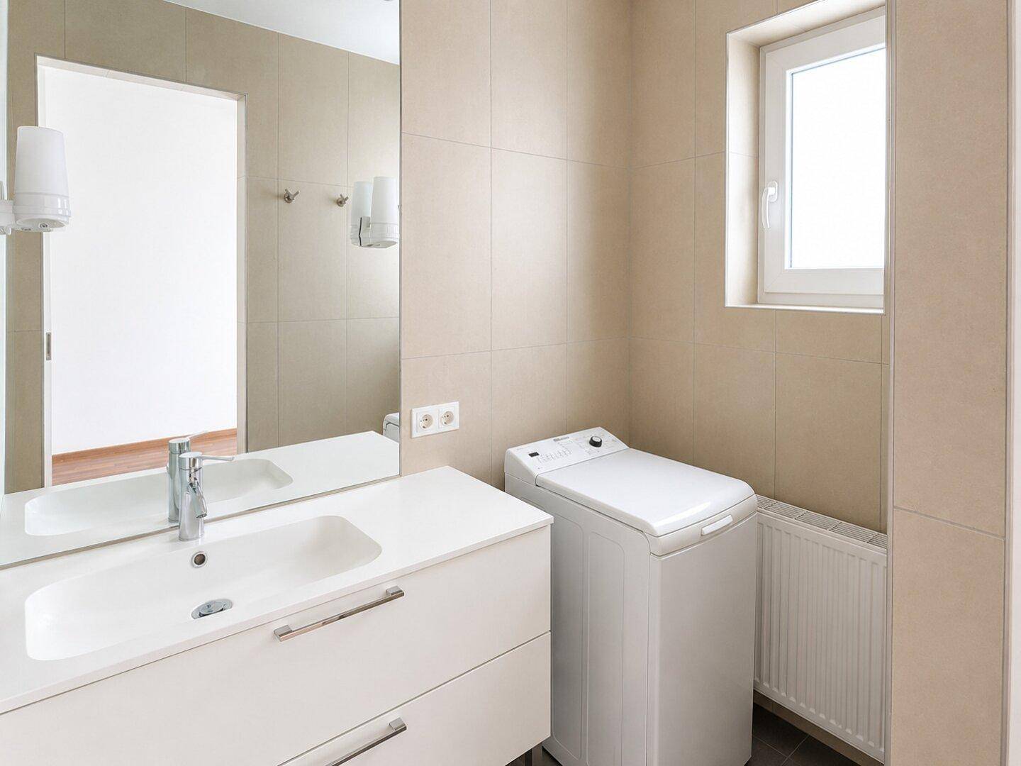 Helles Badezimmer mit Waschbecken, großem Spiegel, Waschmaschine und Fenster für Tageslicht.