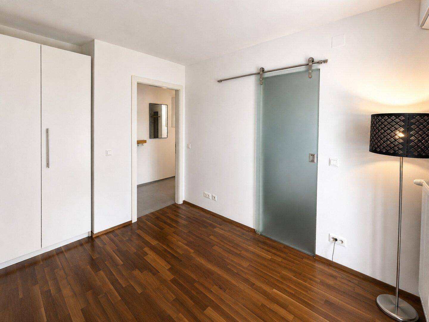 Schlafzimmer mit Holzboden, weißem Einbauschrank und moderner Schiebetür aus Milchglas.