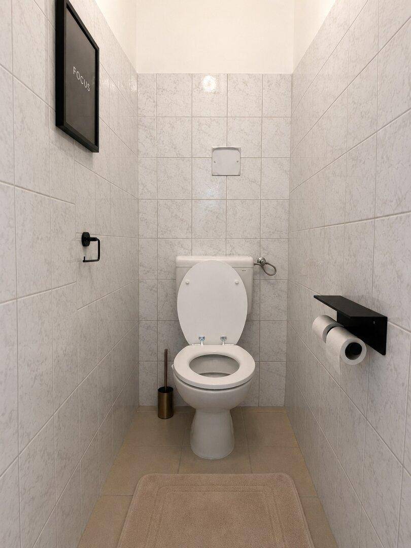 Separates WC mit weißen Fliesen, wandmontierter Toilette und minimalistischer Dekoration.