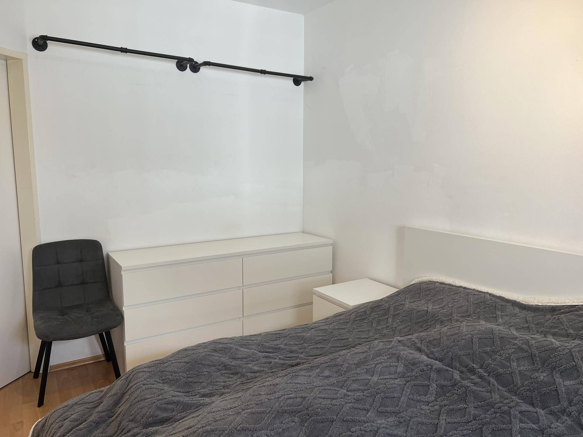 Schlafzimmer mit weißer Kommode, Nachttisch und einem einfachen Kleiderständer an der Wand.