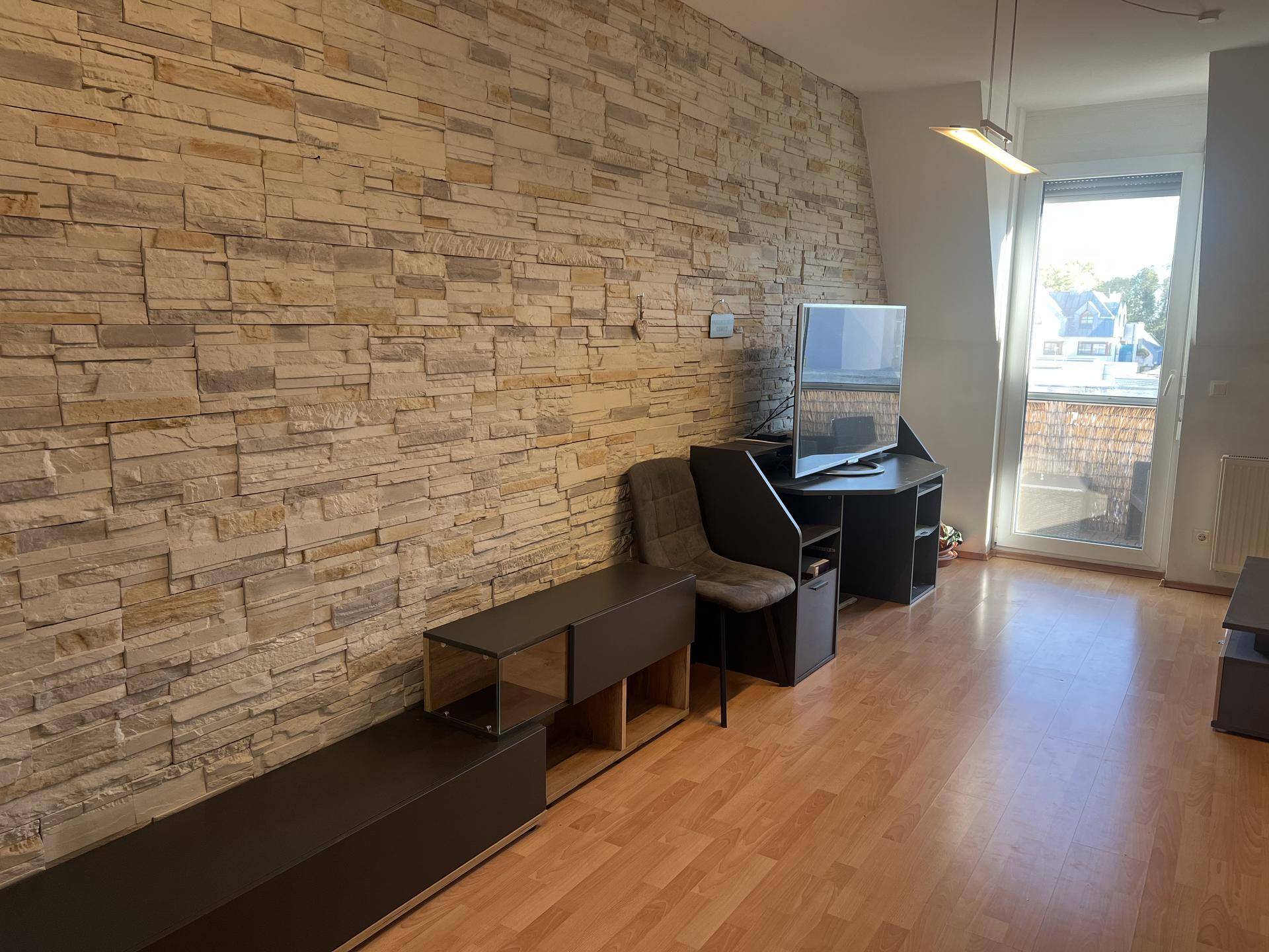 Helles Wohnzimmer mit dekorativer Steinwand, Laminatboden und Zugang zu einem Balkon.