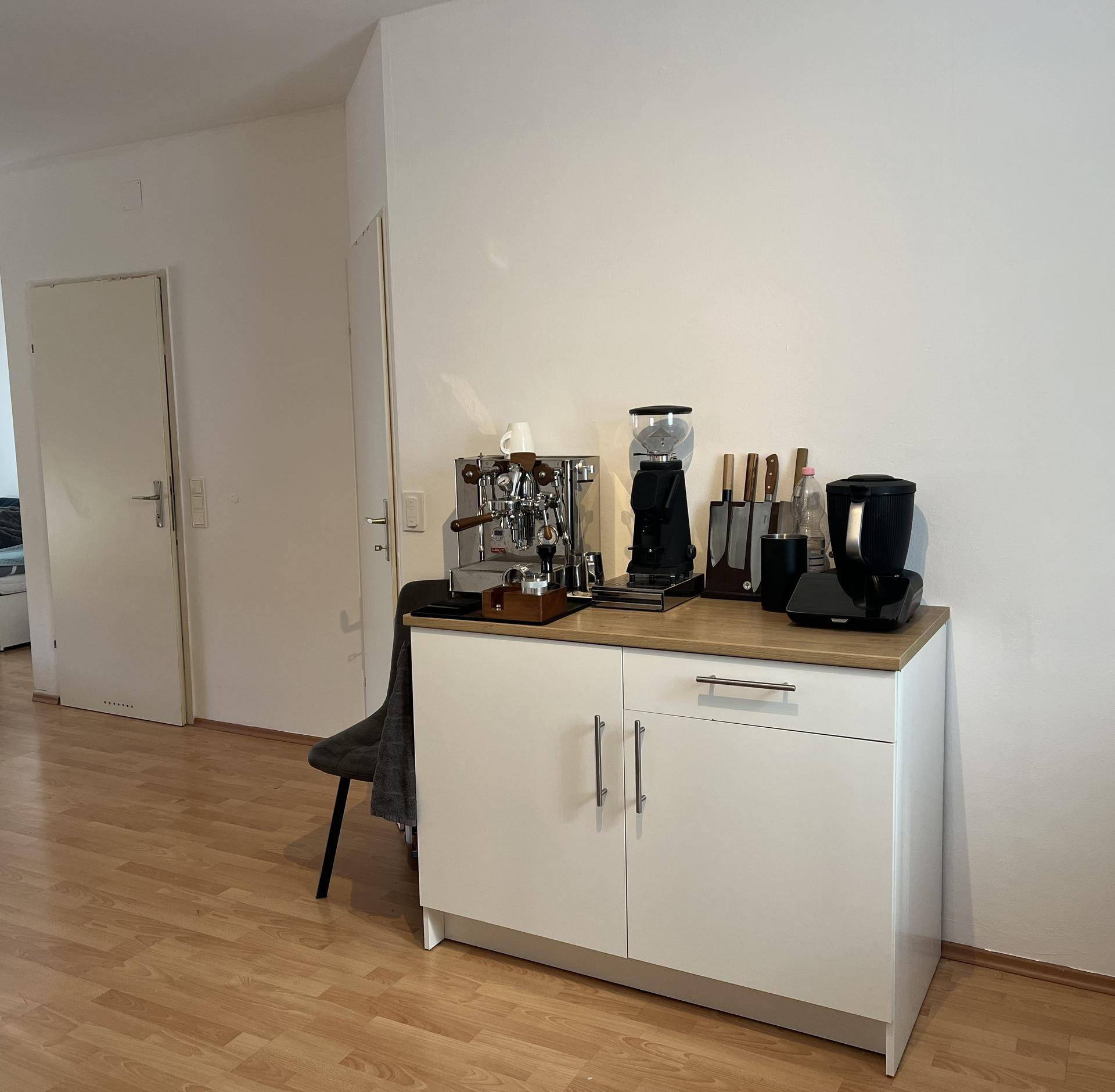 Bereich mit einer Kaffeestation, Schränken und Holzarbeitsplatte in einem hellen Raum.