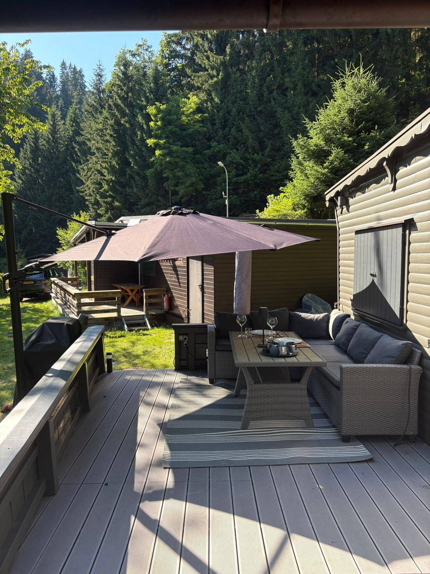 Sonnige Terrasse mit Outdoor-Möbeln und großem Sonnenschirm, umgeben von Wald.