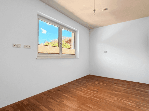 Leeres Zimmer mit hellem Holzboden, weißen Wänden und einem Fenster, das viel Tageslicht hereinlässt.