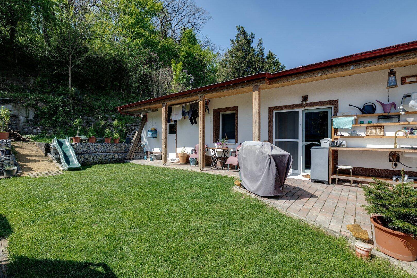 Überdachte Terrasse mit Sitzgelegenheiten und Grillbereich, angrenzend an den grünen Garten mit Rutsche.
