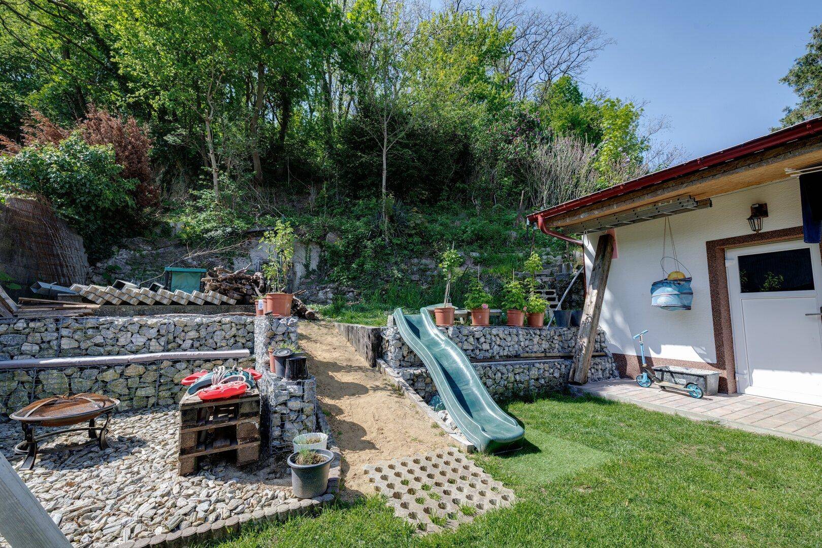 Gestalteter Gartenbereich mit Gabionenmauern, einer Kinderrutsche und verschiedenen Topfpflanzen.
