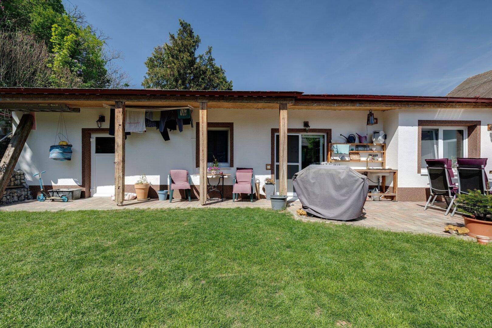 Lange überdachte Terrasse entlang der Hausfassade mit mehreren Sitzbereichen und Grill.